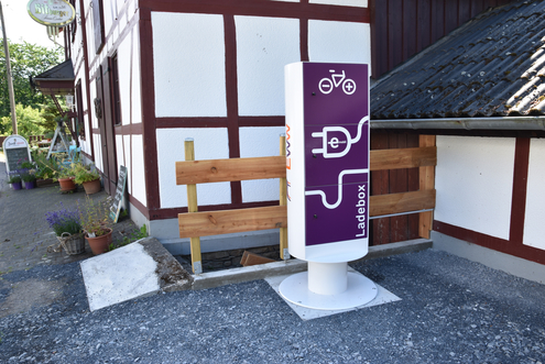 Pedelec-Ladestation_Kraeutercafe Öffentliche Ladesäule für E-Bikes vor einem Fachwerkgebäude.