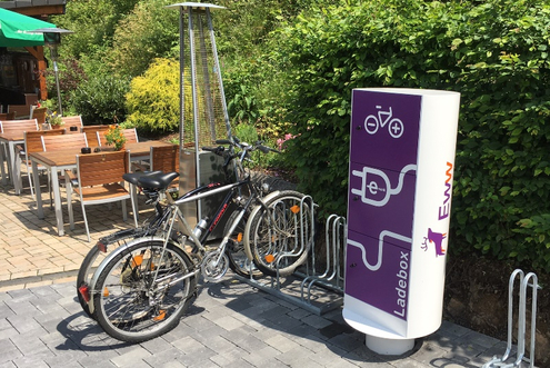 Pedelecbox_Vierbuchermuehle_Websize Öffentliche Ladesäule für E-Bikes mit angeschlossenen Fahrrädern im Außenbereich eines Cafés.
